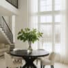 Ronde zwarte eettafel van 150 cm met kruispoot, geplaatst in een licht en modern interieur met hoge plafondvide en grote ramen die veel daglicht binnenlaten. Vier beige stoelen staan rond de tafel en een glazen vaas met groene takken staat centraal op het tafelblad.