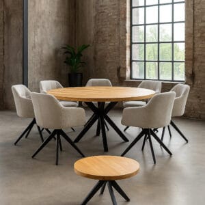 Industriële ruimte met een ronde mangohouten eettafel met licht houten blad en zwarte kruisvormige poten, omringd door acht beige gestoffeerde stoelen. Op de voorgrond staat een bijpassende lage ronde mangohouten salontafel. De ruimte heeft betonnen vloeren, bakstenen muren en een groot metalen raamkozijn.”